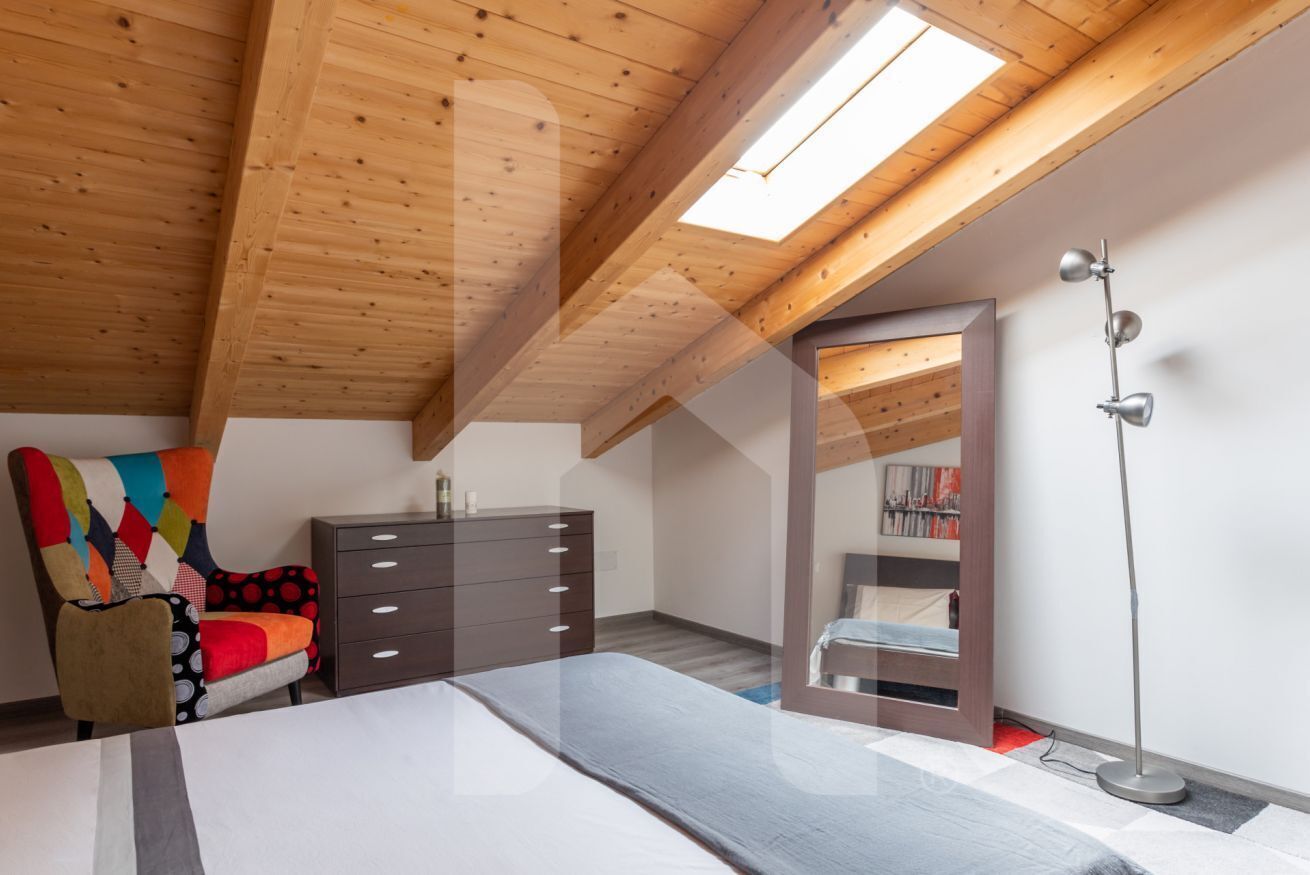 A bedroom with a sloped wooden ceiling, a bed, a dresser, a colorful chair, a floor lamp, and a mirror reflecting a bookshelf.