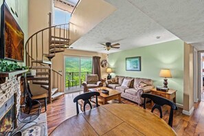 A spacious living area with a wooden dining table, a sofa, and a fireplace. A spiral staircase leads to a balcony with a view of greenery.
