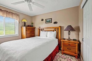 Un dormitorio con cama, mesitas de noche de madera, ventilador de techo, ventana con persianas y una puerta.