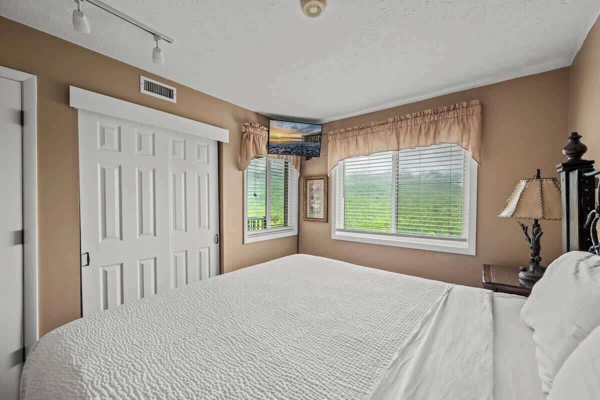 Quarto com uma cama grande, porta, janelas com persianas, um respiradouro no teto e uma TV fixada na parede.