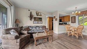A spacious living room with a sofa and coffee table, a dining area with wooden chairs, and a kitchen visible through an open doorway. - Smoky Mountain Condo w/ Heated Pool & Sauna Access (Gatlinburg)