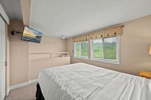 A bedroom with a bed, a TV mounted on the wall, a window with blinds, and a view of greenery outside.