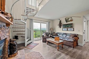 A spacious living room with a stone fireplace, a wooden coffee table, a patterned sofa, and a spiral staircase.