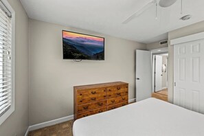 Um quarto com uma cama, uma cômoda de madeira, uma televisão na parede e uma porta que dá para outro espaço.