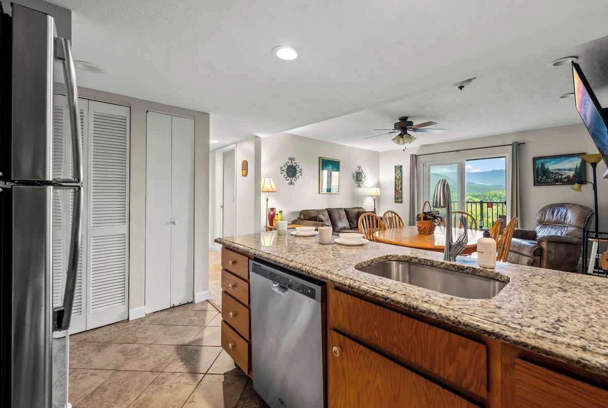 A kitchen with a granite countertop, stainless steel sink, and a dishwasher. There is a dining area with a table and chairs, and a ceiling fan.