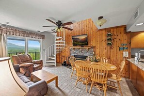 Un salon chaleureux avec une cheminée, des murs en bois, un coin repas et un balcon offrant une vue magnifique.