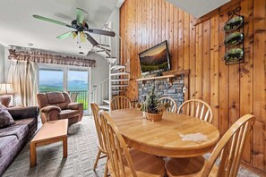 Un salón acogedor con una mesa de comedor de madera, asientos cómodos, una chimenea y vistas panorámicas por las ventanas.