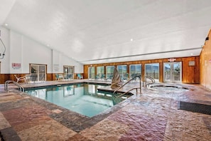An indoor swimming pool with a hot tub, lounge chairs, and a view of the outdoors.