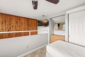 Un dormitorio con techo inclinado, televisor montado, panel de pared de madera, puerta blanca y cama.
