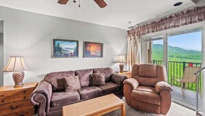 Ein Wohnzimmer mit einem braunen Ledersofa, einem Holztisch und einem braunen Leder-Sessel. An den Wänden hängen zwei Gemälde, es gibt einen Deckenventilator und einen Blick auf grüne H ügel durch große Fenster.