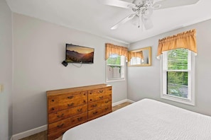 A bedroom with a bed, a wooden dresser, a ceiling fan, a wall-mounted TV, and windows with curtains.