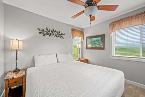Un dormitorio con una cama grande, un ventilador de techo, una mesita de noche con lámpara, una ventana con cortinas y un adorno en la pared.