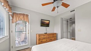 Un dormitorio con cama, cómoda de madera, ventilador de techo, ventana con persianas y puerta que da a otra habitación.