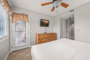 Une chambre à coucher comprenant un lit, une commode en bois, un ventilateur de plafond, une fenêtre avec des stores et une porte donnant sur une autre pièce.