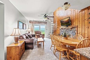 Amplia sala de estar con sofá, sillas, una mesa de comedor y chimenea. Se ve una escalera que conduce a un nivel superior, y un televisor montado en la pared.