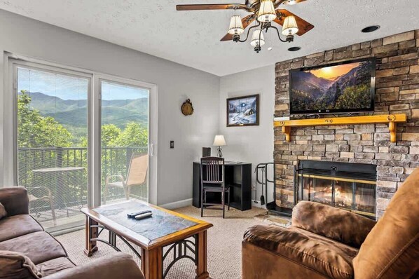 A living room with a fireplace, a television mounted on a stone wall, and a balcony with a view of the mountains.