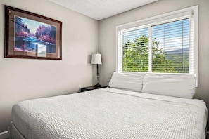 A bedroom with a large bed, a nightstand with a lamp, a framed picture on the wall, and a window with blinds.