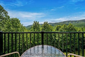 A balcony with a glass table and chairs, overlooking a forested landscape with hills in the distance.