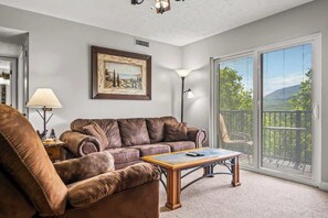A living room with a brown leather sofa, a wooden coffee table, a floor lamp, and a large window with a view of greenery. - Mountain View Balcony, Heated Pool, Near (Gatlinburg)