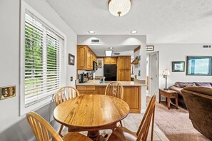 A dining area with a wooden table and chairs, a kitchen with wooden cabinets and a refrigerator, and a living room with a brown sofa and a lamp.