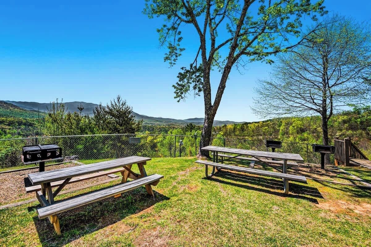 A picnic area with benches and grills, surrounded by a fence and trees, overlooking a hilly landscape.