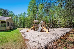 A wooden play structure in a forested area with a covered seating area and a picnic table nearby.