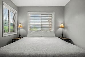 A bedroom with a large bed, two bedside tables with lamps, a window with blinds, and a view of greenery outside.