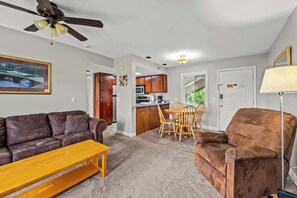 Ein Wohnzimmer mit braunem Ledersofa, einem Holztisch und einem braunen Sessel. Es gibt einen Essbereich mit Holztisch und Stühlen, und im Hintergrund eine Küche.