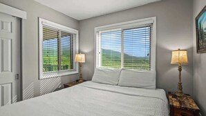 Une chambre à coucher avec un grand lit, deux tables de chevet avec des lampes et deux fenêtres équipées de stores.