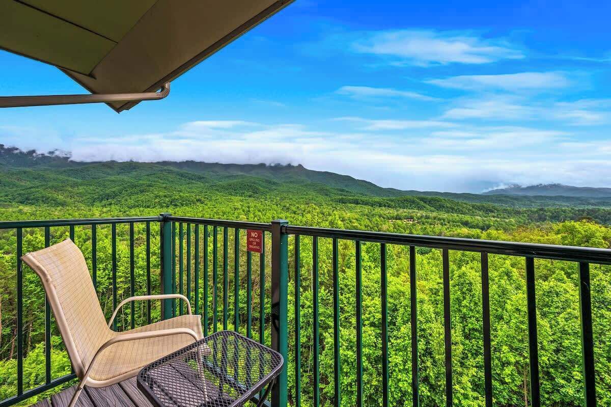 A balcony with a wicker chair and a metal table, overlooking a lush green landscape with distant mountains and a clear blue sky.