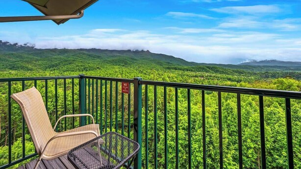 Un balcon agrémenté d’une chaise en osier et d’une table en métal, offrant une vue sur un paysage verdoyant luxuriant, avec des montagnes lointaines et un ciel bleu azur.