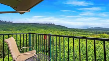 A balcony with a wicker chair and a metal table, overlooking a lush green landscape with distant mountains and a clear blue sky.