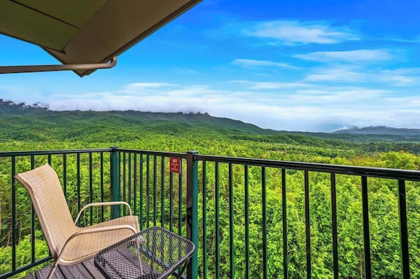 Un balcón con una silla de mimbre y una mesa metálica, con vistas a un paisaje verde exuberante, montañas lejanas y un cielo azul despejado.