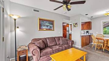 A living room with a brown leather sofa, a wooden coffee table, a floor lamp, and a ceiling fan. There is a kitchen area with wooden cabinets and a dining table with chairs.