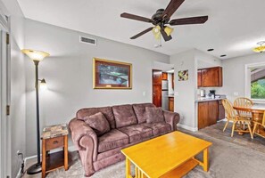 Un salon avec un canapé en cuir marron, une table basse en bois, un lampadaire et un ventilateur de plafond. Il y a également un coin cuisine avec des placards en bois et une table à manger avec des chaises.