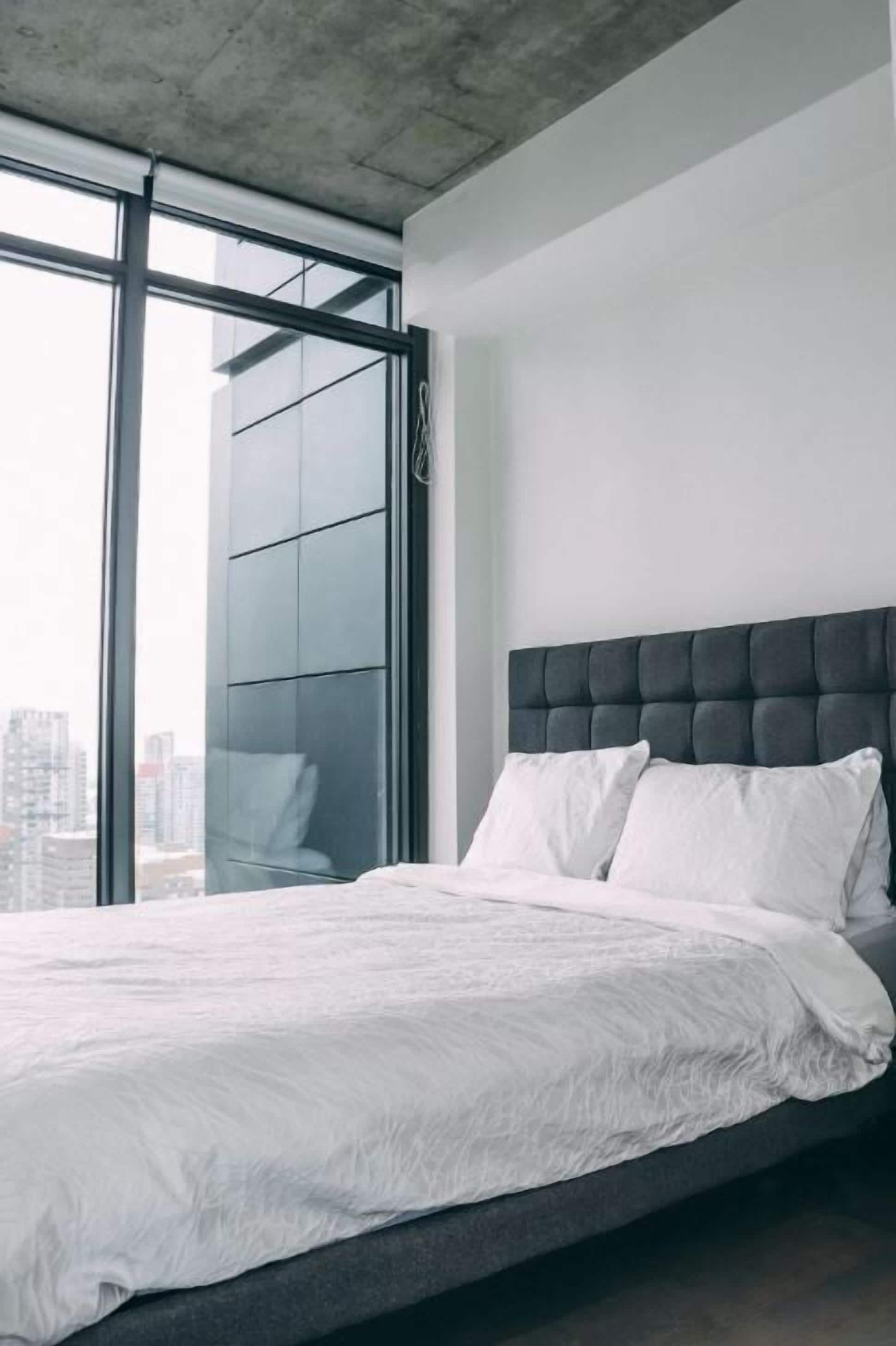 A modern bedroom with a large bed, a grey headboard, and a city view through floor-to-ceiling windows.