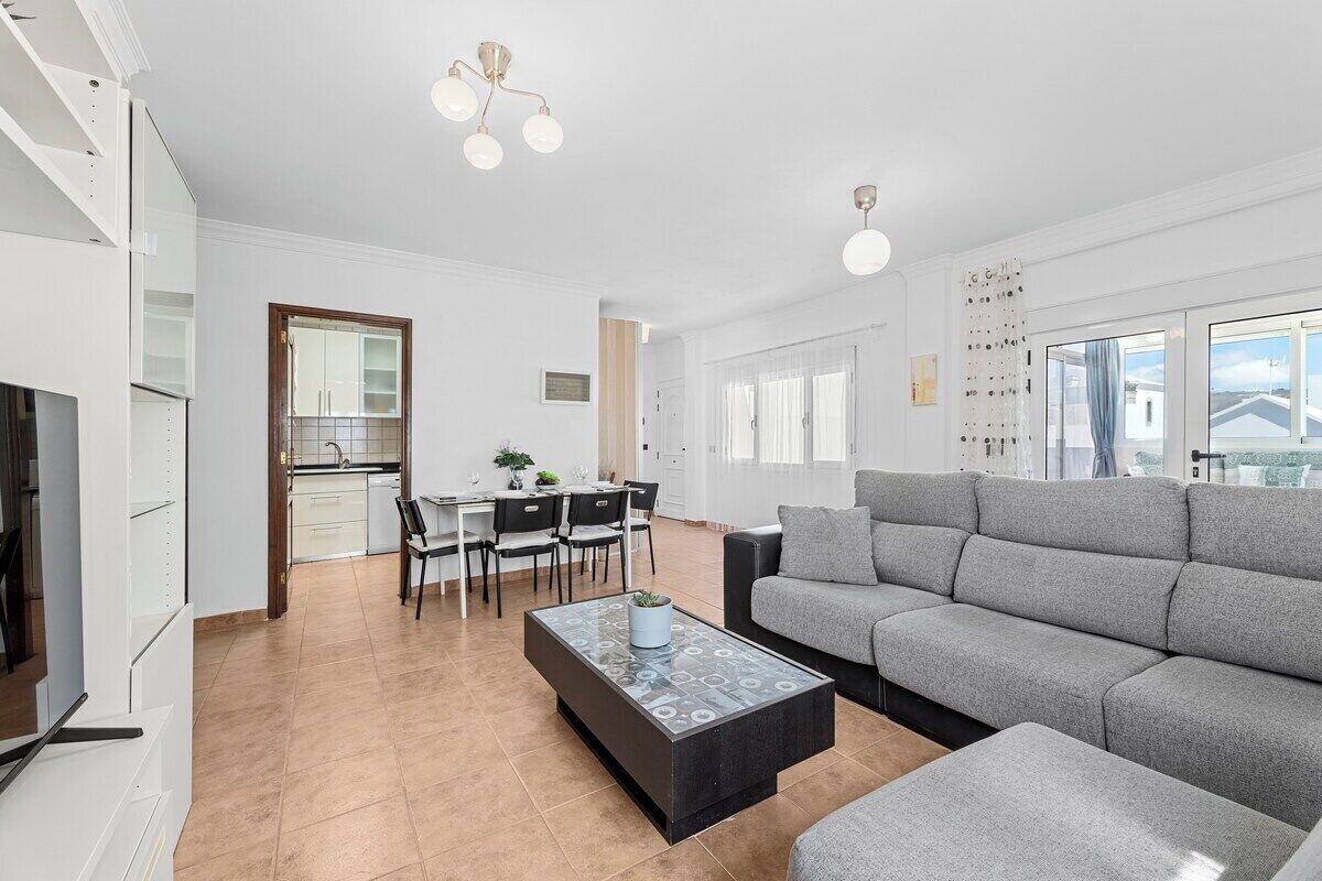 Una sala de estar moderna con un sofá gris, una mesa de centro de vidrio y un comedor con una mesa y sillas blancas.