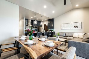 A modern dining area with a wooden table set for four, a kitchen in the background, and a grey sofa in the living area. - Wellness Retreat with Fitness Equipment, Sauna, Cold Plunge and Hot Tub (Kamas)