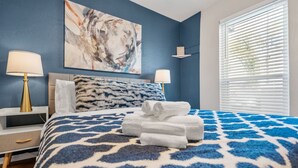 A bedroom with a patterned bedspread, a large abstract painting on the wall, and a window with blinds. - Passionate 2BD: Where Your HEART Feels at HOME! (Fort Worth)