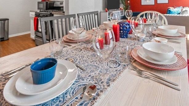 A dining table set with plates, glasses, and candles, with a kitchen in the background. - Passionate 2BD: Where Your HEART Feels at HOME! (Fort Worth)