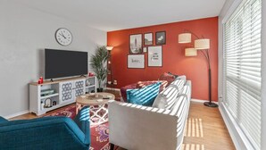 A living room with a TV, sofa, and coffee table. There are framed pictures on the wall and a plant. - Passionate 2BD: Where Your HEART Feels at HOME! (Fort Worth)