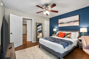 A bedroom with a bed, two bedside tables with lamps, a ceiling fan, a TV, and a door leading to another room. - Vivid 1BD: TCU Walkable, Inspiring & Restful! (Fort Worth)