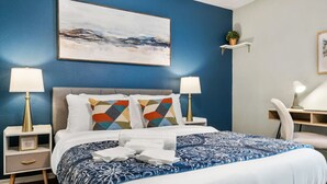 A neatly made bed with a blue and white patterned bedspread, two bedside lamps, and a wall-mounted abstract painting. - Vivid 1BD: TCU Walkable, Inspiring & Restful! (Fort Worth)