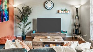 A living room with a large flat-screen TV, a wooden coffee table, a sofa, a floor lamp, and a potted plant. - Vivid 1BD: TCU Walkable, Inspiring & Restful! (Fort Worth)