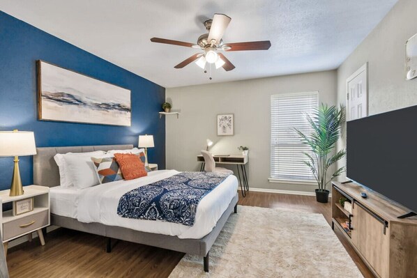 A bedroom with a bed, bedside tables, a ceiling fan, a desk, a chair, a TV, and a window with blinds. - Vivid 1BD: TCU Walkable, Inspiring & Restful! (Fort Worth)