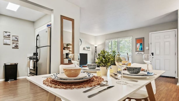 A dining area with a white table set for two, a refrigerator, a trash can, and a window with blinds. - Vivid 1BD: TCU Walkable, Inspiring & Restful! (Fort Worth)