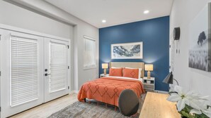 Une chambre à coucher comprenant un lit, deux tables de chevet avec des lampes, une grande fenêtre avec des stores et une œuvre d’art accrochée au mur.