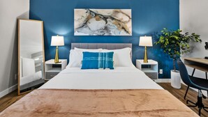 Una habitación de hotel moderna con una cama grande, dos mesitas de noche con lámparas, un escritorio y un cuadro abstracto de gran tamaño en la pared.