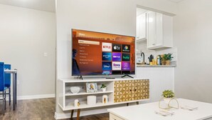 Uma sala de estar moderna com uma TV de tela plana exibindo a interface de um serviço de streaming, um móvel branco com prateleiras, uma mesa de centro e um espaço de refeições com uma cadeira azul.