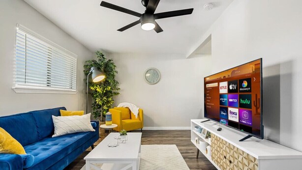 Uma sala de estar moderna com um sofá azul, uma mesa de centro branca e uma TV de tela plana exibindo serviços de streaming.